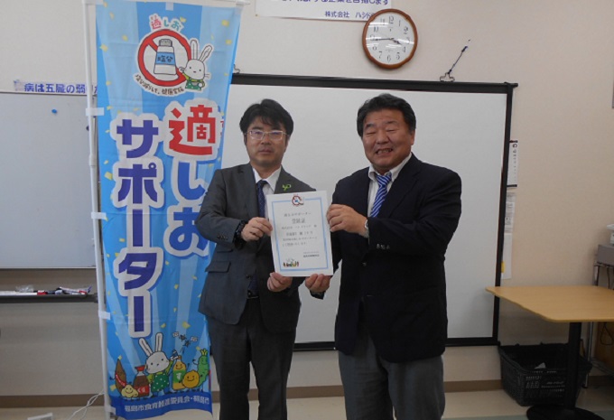 「適しおサポーター」の水色ののぼりの横に立ったスーツ姿の男性二人が両側から登録証を持った記念写真