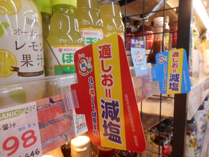 レモン果汁などが並んだ陳列棚に「適しお＝減塩」と大きく描かれた赤と黄色、水色と黄色の配色のポップが取り付けられている写真