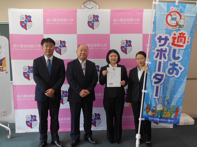 桜の聖母短期大学のバックボード前にスーツを着た男性2人と登録証を胸の前で持った女性、「適しおサポーター」の青いのぼりを持った女性が並んで立っている写真