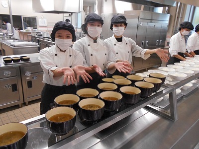 厨房でコック服と帽子、マスクを身に着けた女性3人が、台に並んだ汁が入ったたくさんの汁椀を手で示している写真