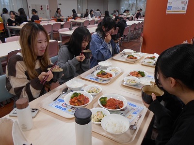 カフェテリアのテーブル席に着いた女性たちが、それぞれの前に置かれた料理の乗ったトレーから汁椀をとり、おいしそうに食べている写真