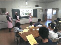 長机の席に座った子供たちの前にピンクの三角巾とエプロンを身に着け、マスクをつけた女性が3人並んで立っている写真
