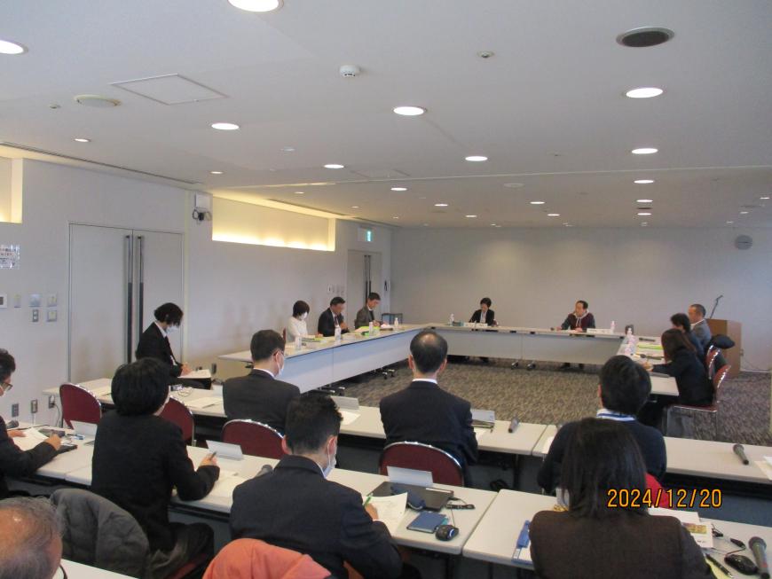 会議室にコの字型に並んだ会議机とその正面に横に数列並んだ会議机の席に出席者たちが座っている委員会の様子の写真