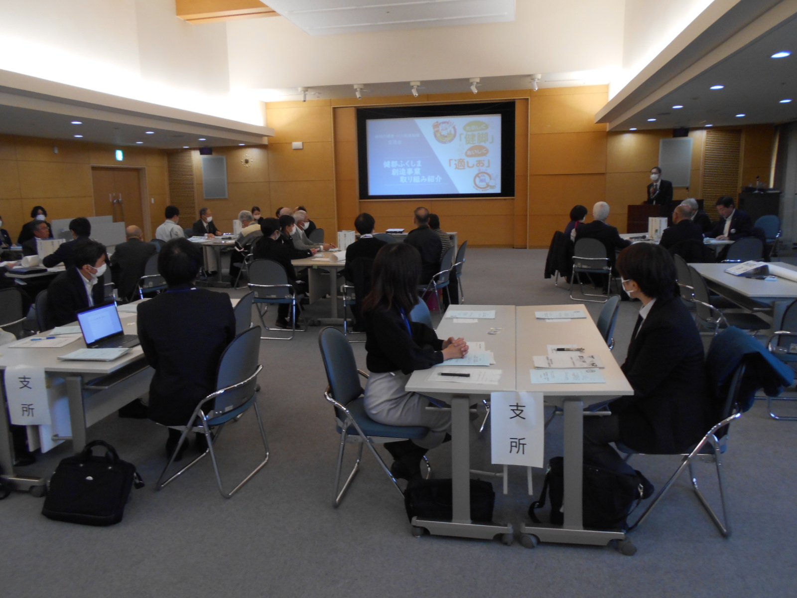奥の壁に大きなスクリーンがあり、グループに分かれて大勢の参加者がテーブルの席に座っている会場全体の写真