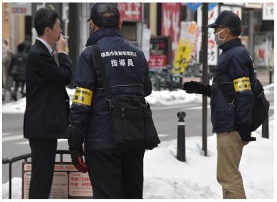 路上で喫煙している男性に指導員の上着を着て腕章をつけた男性2人が注意・指導をしている写真