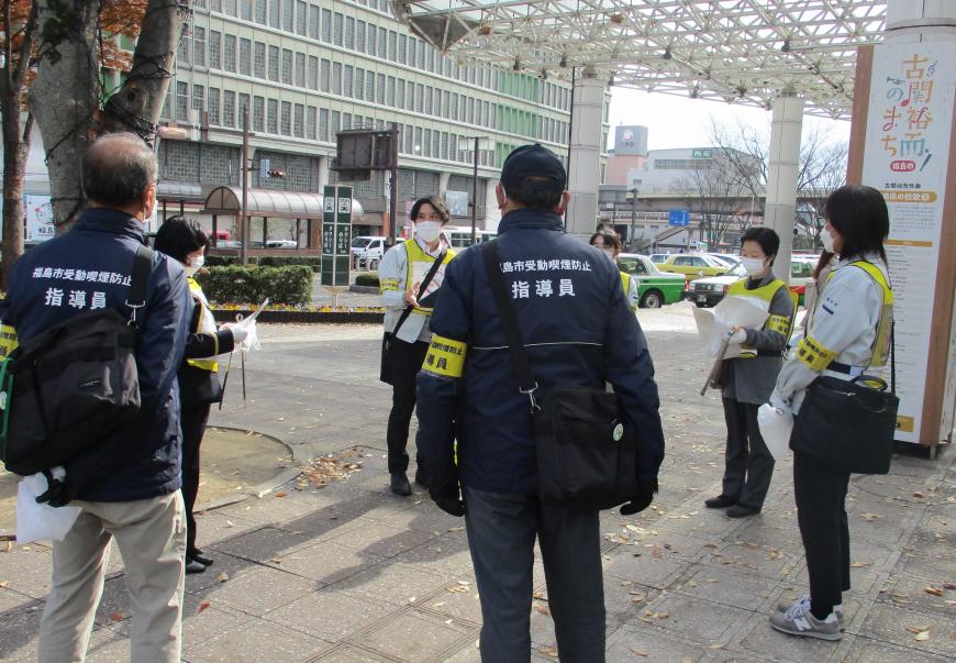 駅前のスペースで、黄色のビブスや腕章を身に着けた参加者たちが輪になって話をしている様子の写真