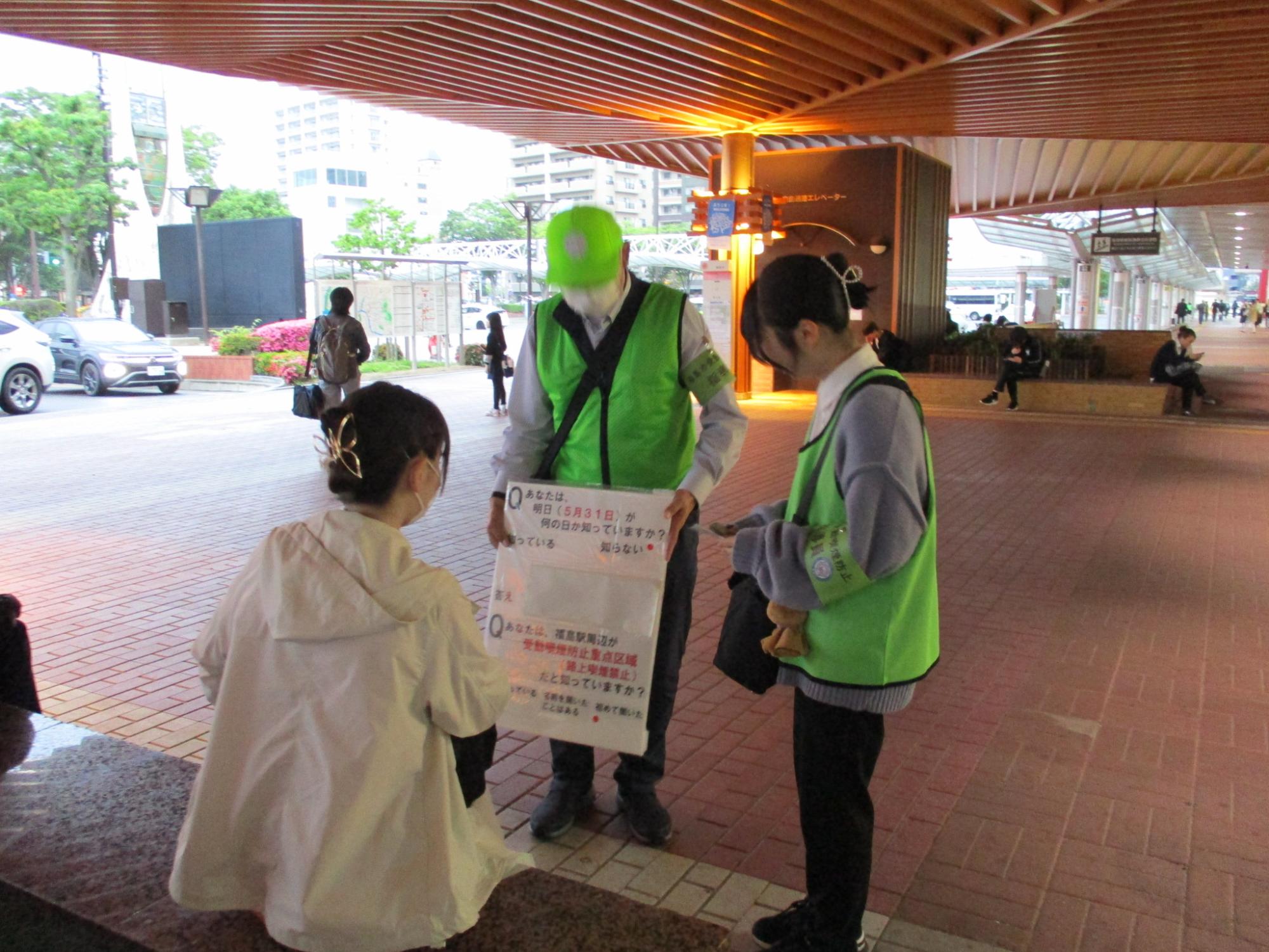 黄緑色のビブスを着た男女が駅前にいた人に街頭アンケートに答えてもらっている写真