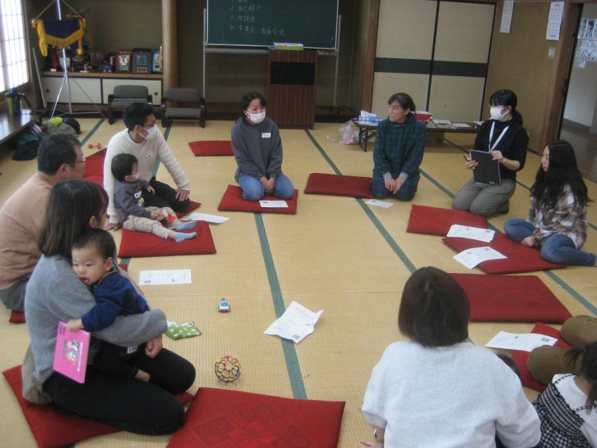 座敷に赤い座布団が丸く並べられ、小さい子供を連れた参加者たちが輪になって話をしている写真