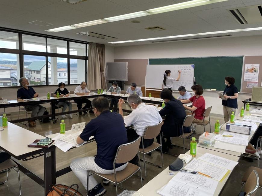 口の字型に並べられた長机の席に協議会役員が座り、黒板の前に置かれたホワイトボードに女性が書き込んでいる協議会の様子の写真