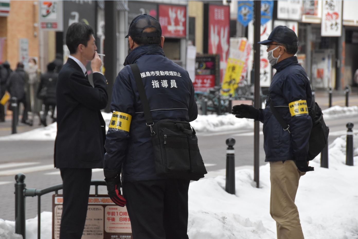 黄色い腕章をつけた男性2人が歩道で喫煙している男性に声をかけている写真
