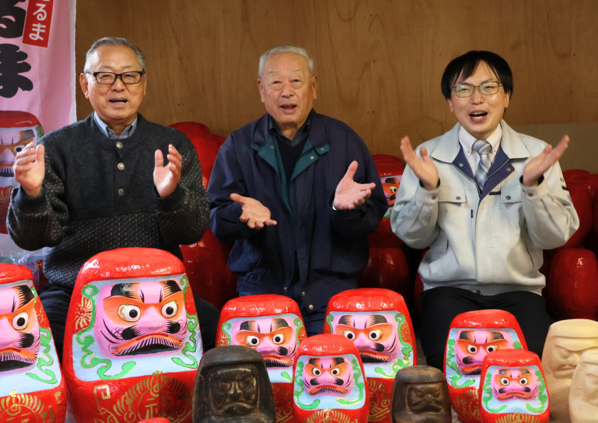 ダルマに囲まれた高野会長と村田さんと安田さんが両手を広げ並んで座っている写真