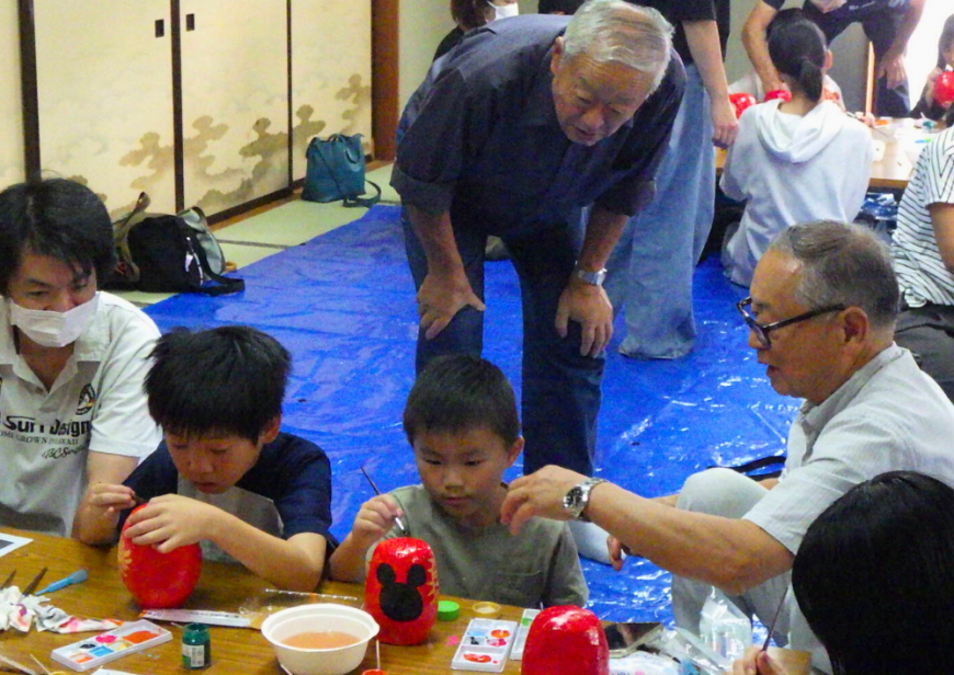 傍で父親や2名の男性が見守るなか、2人の男の子がダルマに色付けしている様子の写真