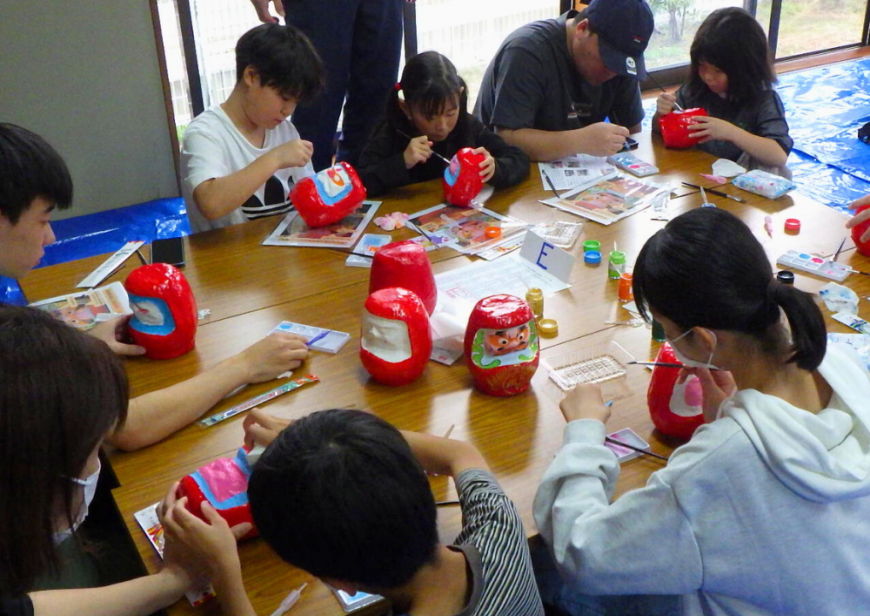 3台の長机をくっつけた周りに参加した親子連れが座り、ダルマの絵付け体験を行っている様子の写真