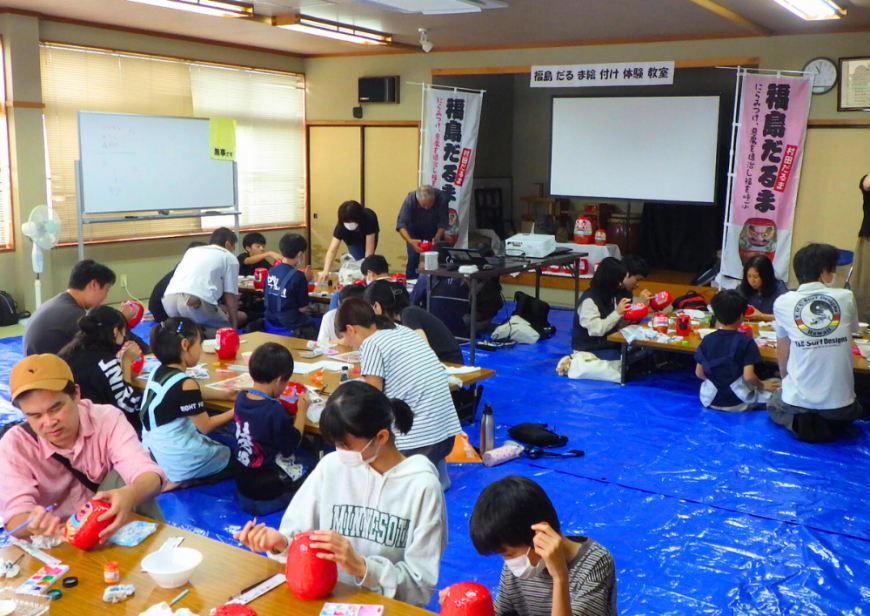 床にビニールシートが敷かれた屋内で机ごとに分かれて座っている参加者たちが、だるま作り体験を行っている様子の写真