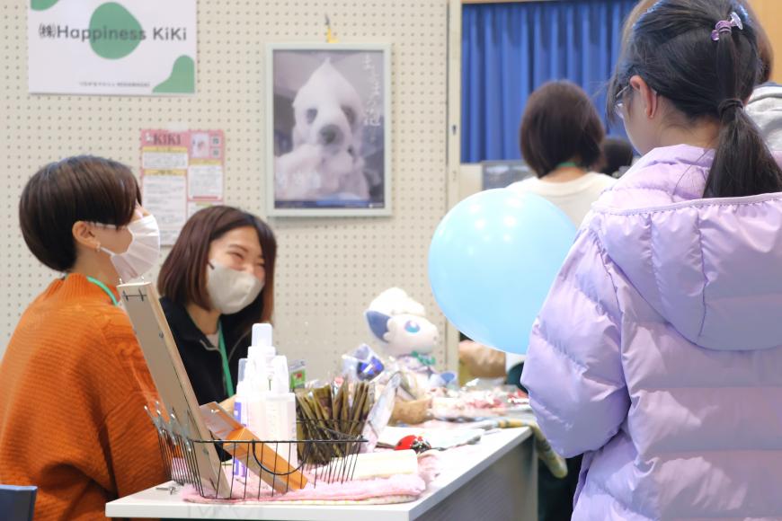 イベントブースの机で笑顔で接客をする女性と来場者が写っている「つながるマルシェMIKAWADAI」のイベントの様子の写真