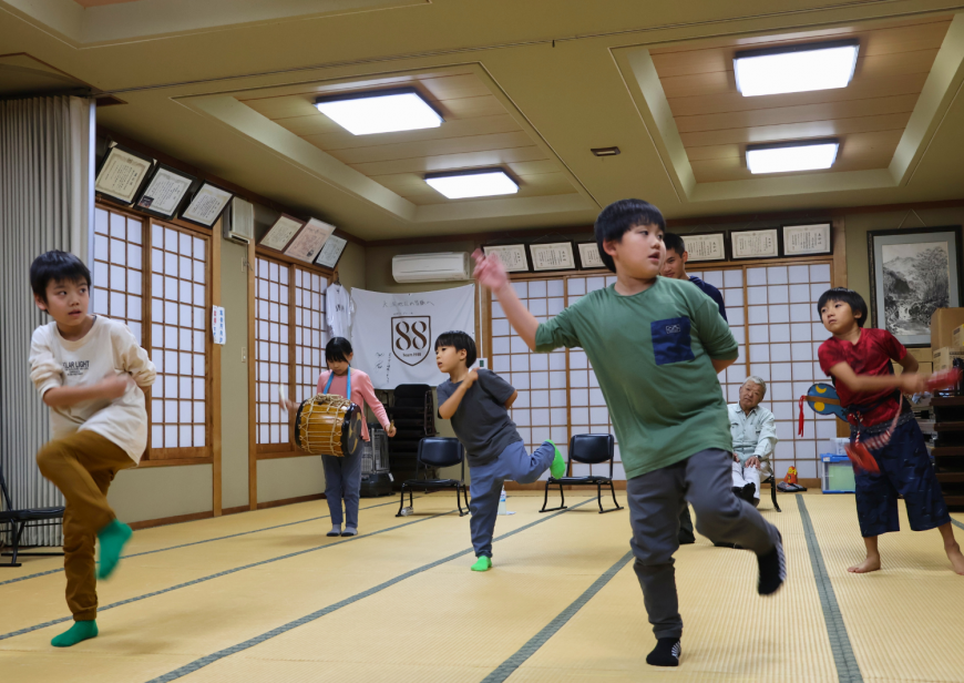 間隔をあけて男子児童が並び、左足を上げ、左手を背中に右手を胸の前に置き踊りの練習を行っている写真
