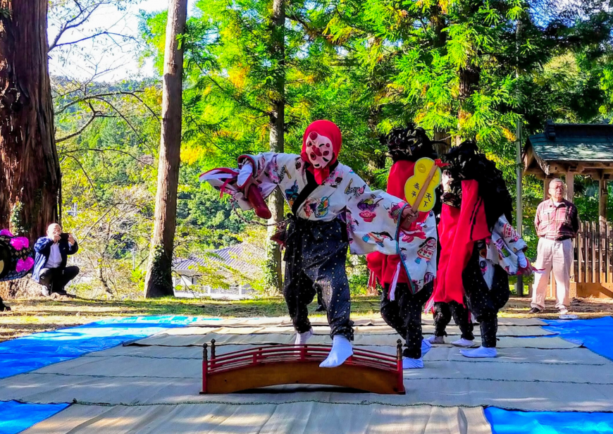 神社の境内に敷かれたビニールシートの上で、頭に赤い頭巾を被りお面をつけ、白地に柄入りの着物とドット柄の紺色のモンペの様な衣装を身にまとった演者が小さな橋の上を跨いで踊っている写真