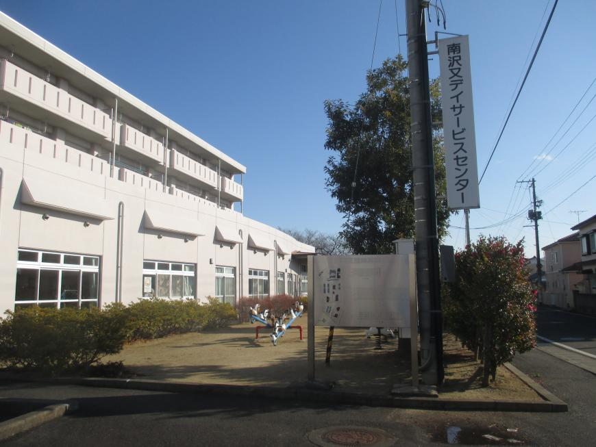 電柱に「南沢又デイサービスセンター」と書かれた看板があり、左側に白い3階建ての大きな建物が写っている写真