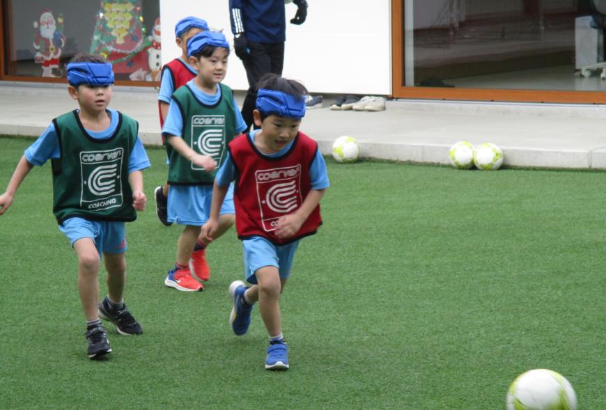 ヘッドガードをし、緑や赤のベストを着用した子どもたちが芝生の上でサッカーボールを追いかけて走っている様子の写真