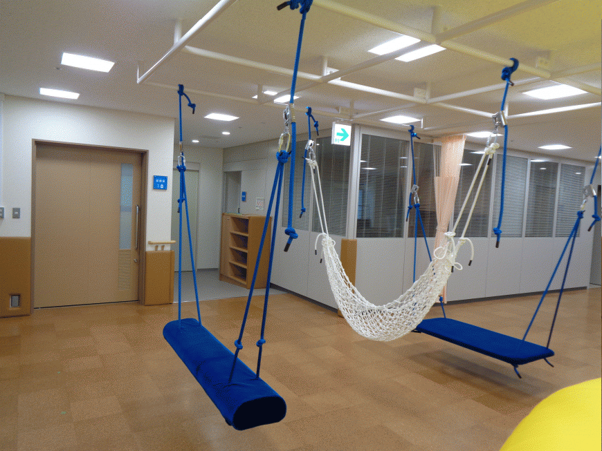 天井から白色のハンモックと、青色のロープで吊り下げられた長方形の道具が設置されたこども発達支援センター室内の写真
