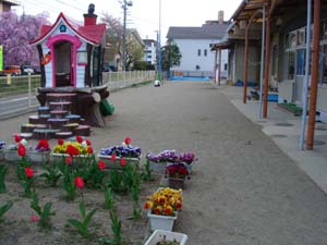 園舎の前に家型の遊具や色とりどりの花が咲いたプランター、チューリップが咲いた花壇があるふくしま中央認定こども園の写真