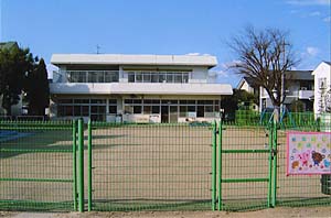 緑色のフェンスの中に広い土の広場が広がり、2階建ての白い建物がたっている御山保育所の外観写真