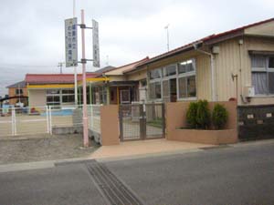 薄茶色の門構えで白いフェンスで囲まれた中に園庭と赤い屋根の1階建ての建物がある渡利保育所の外観写真