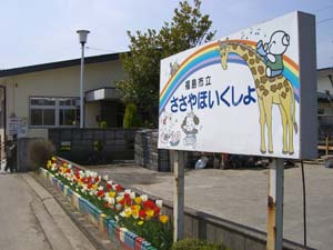 「福島市立 ささやほいくしょ」と書かれた虹や動物が描かれた白い看板の下にチューリップの花壇があり、その先に白い壁の建物がある笹谷保育所の外観写真