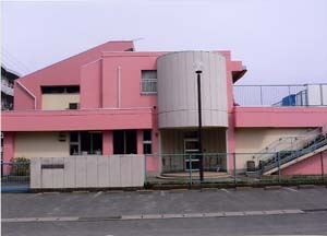 ピンク色と白の外壁の2階建ての建物の前に門と緑色のフェンスがあり、その手前に駐車場がある野田保育所の外観写真