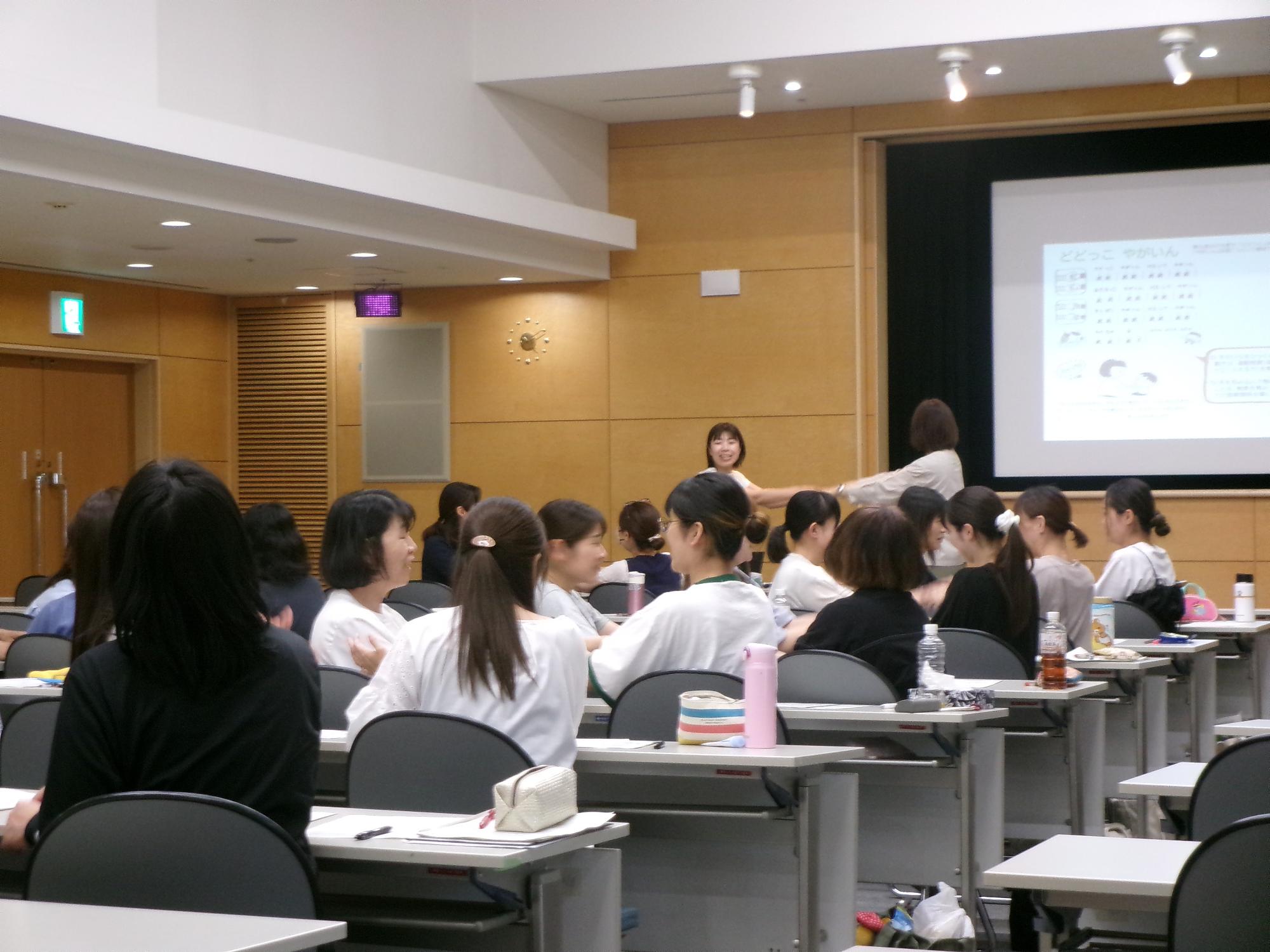 「こどもの発達と遊び」の講義の中で、2人組で向かい合って手をつないで遊ぶ実践をする講師と受講者の写真