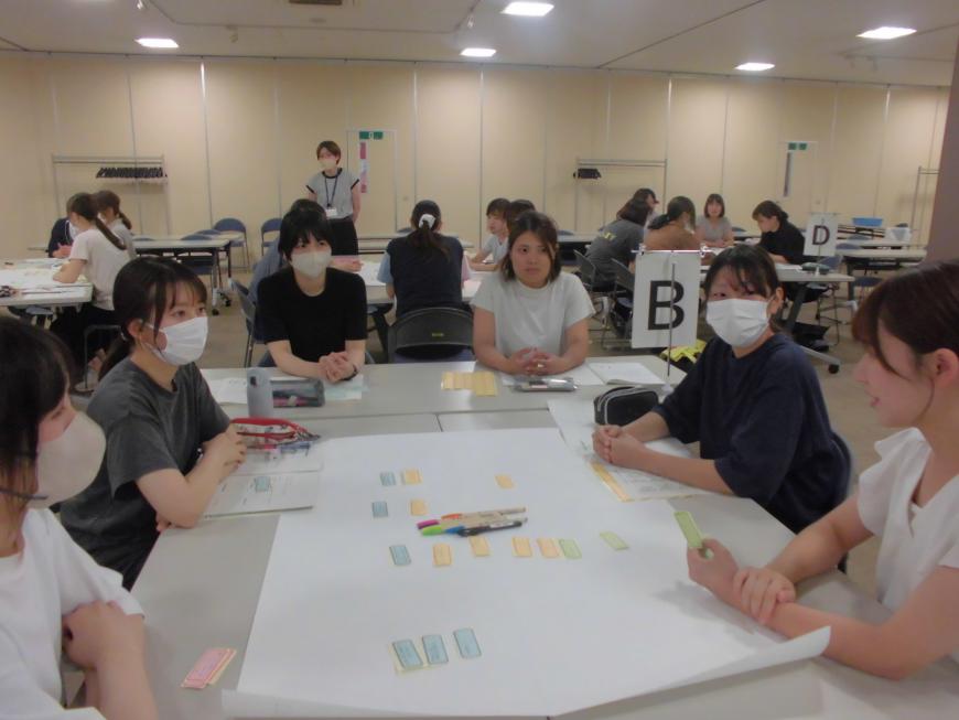 黄色や水色のメモが貼られた模造紙が広げられたテーブルを囲んで6人の女性が座っている写真