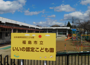 黒いフェンスの前に、「福島市立いいの認定こども園」と書かれた黄色の看板が設置され、その後ろには遊具の並んだ園庭、L字型の平屋の園舎がある、いいの認定こども園の外観写真