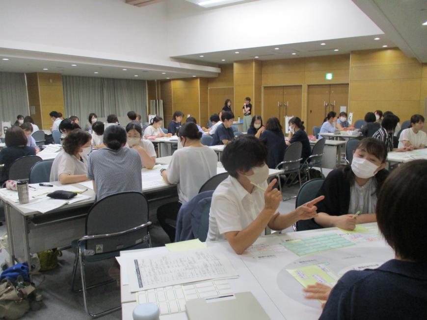 グループに分かれて席に座ったたくさんの出席者がテーブルに資料を広げて話し合いをしている写真