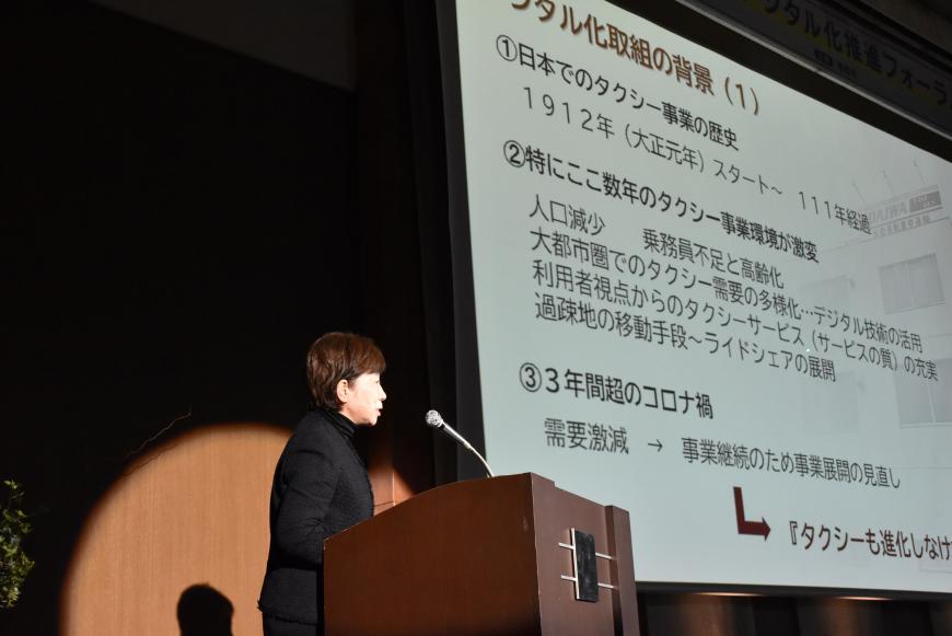資料が映し出されたスクリーンの左側に設置された壇上に立つ大村氏が話をしている様子の写真