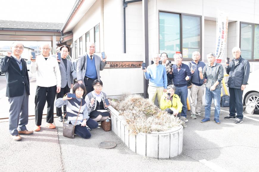 シルバー人材センターの看板が設置された入り口に、それぞれスマートフォンを持った皆さんが並んでいる写真