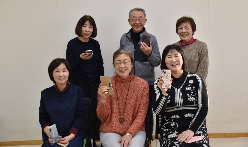前列の椅子に3人、後列に3人のメンバーがそれぞれスマートフォンを持ち並んでいる写真