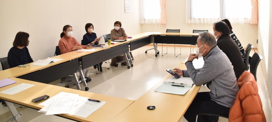参加者がロの字型に設置した長机の席に座り、中央に座っている女性の話を聞いている写真