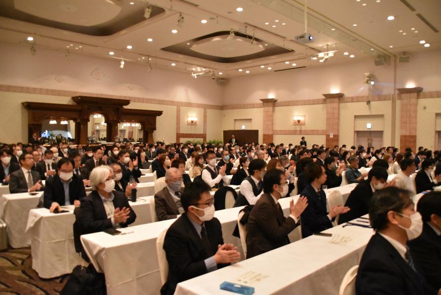 白いクロスが掛けられた机の席に座った参加者たちが拍手をおくっている写真