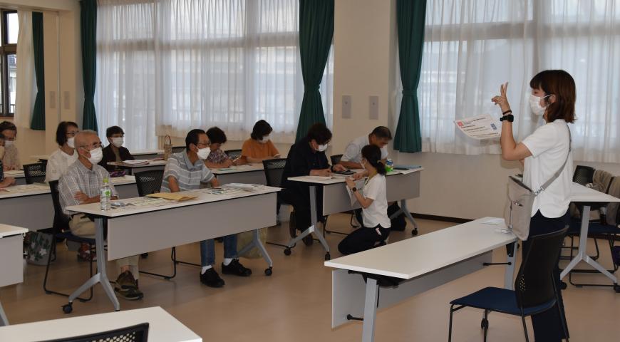 前方に立つ女性講師が話しているのを、長机の席に2名ずつ座った参加者たちが聞いている講座の様子の写真
