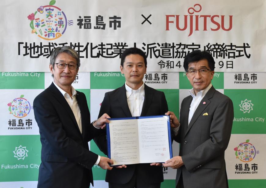 福島市のバックパネルの前で協定書を開いた状態で中央の男性が持ち、市長と関係者の男性が一緒に持って並んでいる写真