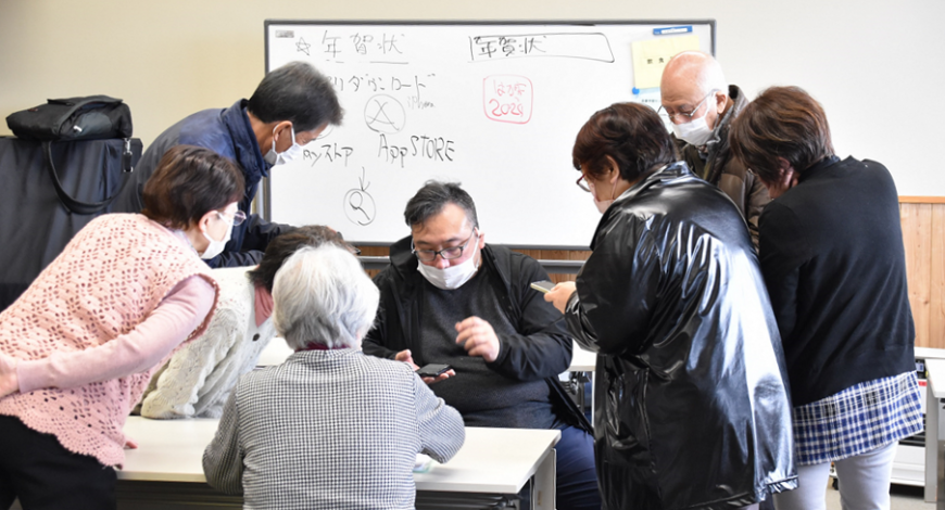 ホワイトボードの前に座りスマートフォンを操作している佐藤講師の周りに参加者が集まっている講習の様子の写真
