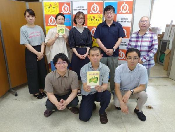 前列に3人、後列に5名立ち、前列中央の男性と後列の左から2番目の女性が冊子を持って並んで写っている集合写真