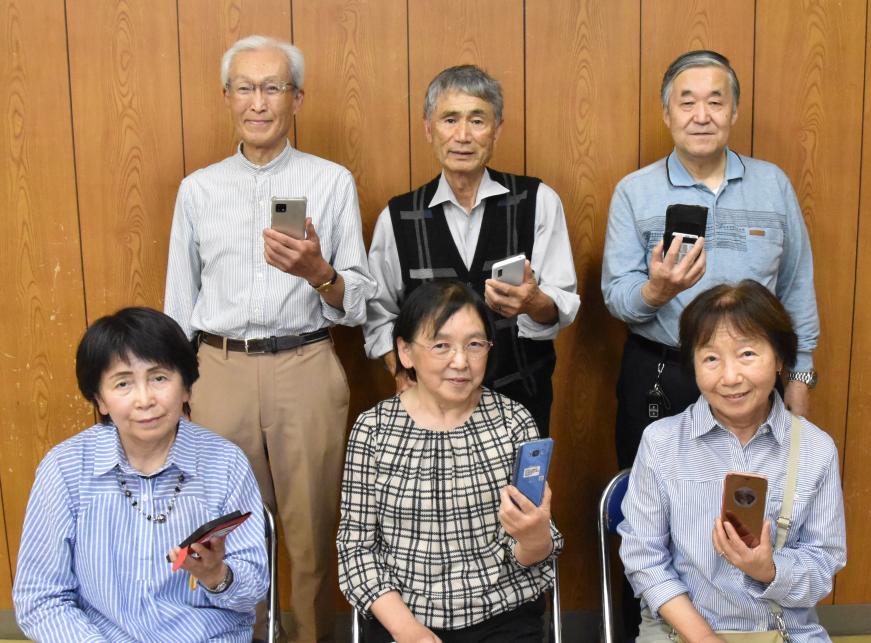 前列の椅子に女性が3人、後列に男性3人のメンバーがそれぞれスマートフォンを持ち並んでいる写真