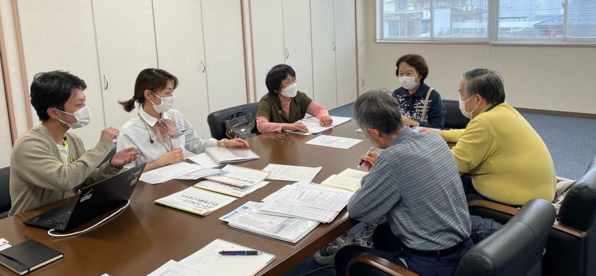 資料やパソコンが置かれた大きなテーブルの席に携帯ショップの担当者やメンバーが座り話をしている写真