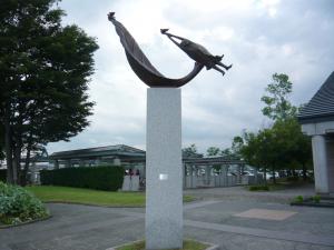 大きな白い柱の上に「し」の字型の様な形の右側に人が両手を伸ばして飛んでいる様を表した作品「大空へ」の写真