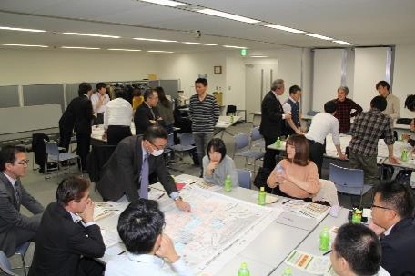 机に資料を広げ、1人の男性が資料を指さしながら周りの参加者の人たちに説明をしている写真