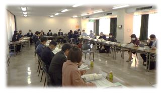 縦2列に並んだ長テーブルと後方に横1列に並べた長テーブルの前に数十名の関係者が着席し、資料を見ながら話をしている会議の様子の写真