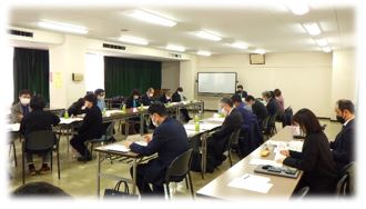 長テーブルを会場内一面に設置し、その前に着席した関係者が資料を眺めたり、話し合ったりしている様子の写真