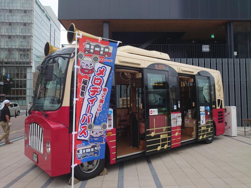 車両の前に「メロディーバス」と書かれたのぼり旗が立っている赤とクリーム色のツートンカラーのあかメロディのバスが建物前に駐車されている写真