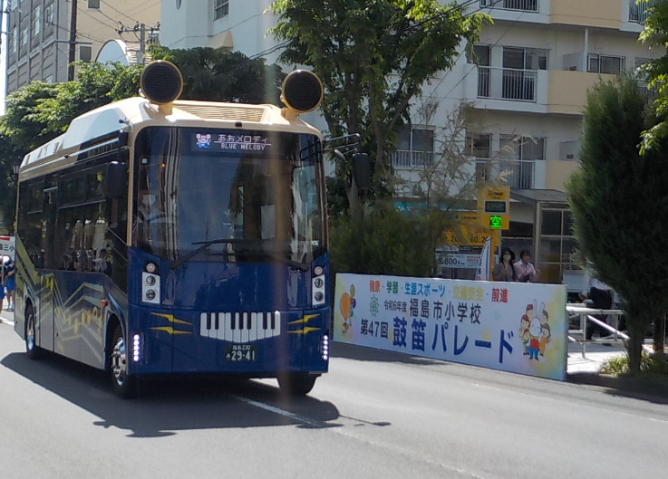 「鼓笛パレード」と書かれた看板の前を走っている車両の頭上にラッパ型のスピーカーが設置された青色のあおメロディバスの写真
